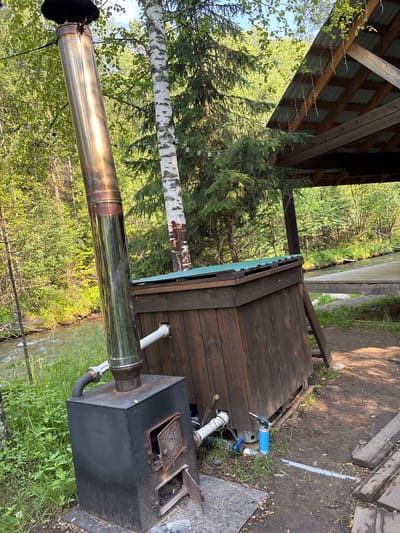 In Bol'shaya Rechka, Russia, an outdoor scene is captured featuring a rustic wooden structure beside a body of water, likely a river. Dominating the foreground is a black metal stove with a tall, shiny stainless steel chimney. The stove has an open firebox, revealing embers and ash. Attached to the stove is a curved pipe leading to a large, weathered wooden tank or hot tub. This tank is partially covered by a green corrugated metal roof. To the right of the tank, a blue gas torch is visible on the ground. Behind the tank, a wooden canopy with a corrugated metal roof provides shelter, supported by a wooden beam. The environment is lush and green, with a forest of birch and pine trees surrounding the area. A river flows gently in the background, reflecting the sunlight. The ground is a mixture of dirt and scattered debris, suggesting a natural, somewhat unkempt setting.