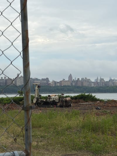 The image depicts an outdoor scene with a city skyline in the background, partially obscured by trees and a body of water. In the foreground, a chain-link fence runs horizontally across the bottom of the frame. To the right of the water and behind the fence, there is a patch of overgrown green grass and some dry brush.
On the right side of the image, a piece of heavy machinery, possibly a drilling rig or a construction vehicle, is parked on a patch of dirt and debris. The machine appears to be white and black with various components and a boom. Next to it, there is a cardboard box and what looks like a stack of cylindrical objects.
The skyline in the background features numerous buildings, suggesting an urban environment. The sky is overcast, with soft, diffused light, indicating it is likely daytime, perhaps late morning or early afternoon, under cloudy conditions. The overall impression is of an industrial or construction site located near a city and a body of water. There are no people visible in the image. No visible text is present.
