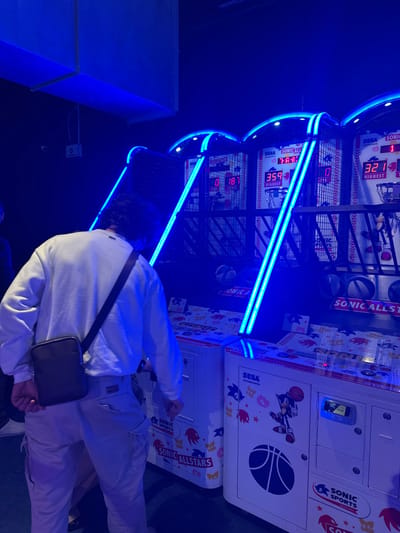 The image displays an indoor entertainment venue in Saint-André-lez-Lille, France, illuminated predominantly by blue ambient light. One individual is centrally positioned, viewed from the back, standing in front of a row of three "Sonic Allstars" basketball arcade games. This individual is a male with dark, medium-length curly hair, wearing a white long-sleeved crew-neck shirt, light-colored cargo pants, and a dark brown or black cross-body bag over his right shoulder. His hands are lowered towards the control panel area of the arcade machine directly in front of him.
The arcade machines are characterized by neon blue illuminated archways and vertical light strips. Each machine displays "SEGA" branding, "SONIC ALLSTARS" logos, and graphics featuring Sonic the Hedgehog playing basketball. Digital score displays show various numbers: "0 18" on the leftmost visible machine, "359 HIGHEST" on the middle machine, and "321 HIGHEST" on the rightmost machine. Small basketballs are present within the collection areas of the games, and a sticker indicating "£ 1.50" is visible near a payment slot on one machine.
The environment is dark, with a high ceiling from which a large, light-colored ventilation duct or structural element protrudes. A small white electrical box or sensor is visible on the dark wall to the upper left. A second individual is partially visible on the far left of the frame, mostly obscured by the main subject. The floor is dark and reflects some of the blue light. The scene depicts active engagement with arcade games in a recreational setting.