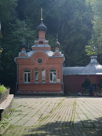 A red brick Orthodox chapel with a dark dome and a golden cross stands in a courtyard paved with rectangular stones, with grass growing between them. The chapel has a series of arched windows on the main facade and a smaller circular window above. To the right of the chapel is a smaller, darker red structure with a silver, multi-faceted roof, which appears to be an outdoor pavilion or covered area. A person in a dark blue shirt is standing inside this structure, near a railing. The background is dominated by dense green trees, suggesting a location with significant natural vegetation. The lighting indicates it is daytime, with sunlight casting shadows on the paved courtyard. This scene is located in the vicinity of the Palikavicki Sel'ski Savet in Belarus.