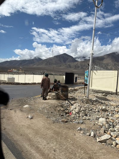 In Phey, India, a scene unfolds with a backdrop of majestic mountains under a bright, cloud-dappled sky. A paved road runs horizontally, bordered by a rocky, uneven terrain. On the roadside, three individuals in military uniforms are present. Two are seated, with one standing and facing away from the camera. Behind them, a light-colored wall topped with barbed wire delineates a perimeter. Beyond the wall, a cluster of buildings, including one with a prominent radio tower, stands against the mountain range. A tall utility pole with electrical wires extends upwards into the sky, its presence marked by a small "Danger" sign. Several dark barrels and scattered rocks are visible in the foreground, adding to the rugged, frontier-like atmosphere.
