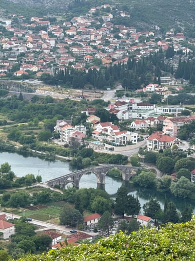 This is a high-angle, vertically oriented aerial shot of a town nestled alongside a river, with a steep, tree-covered mountainside on the left. The town features densely packed houses with red-tiled roofs and white or light-colored walls, some of which are modern with large windows and balconies. The houses are built on a hillside, creating a terraced effect.
A river flows through the scene, spanned by an old stone bridge with multiple arches. The water appears calm and somewhat reflective. Roads and pathways wind through the town and along the riverbanks, with several cars visible, suggesting ongoing activity. Lush green trees and vegetation are abundant throughout the town and on the surrounding slopes, indicating a possibly temperate or Mediterranean climate.
The lighting suggests it is daytime, likely overcast or late afternoon, as there are no harsh shadows. The overall atmosphere is serene and picturesque, showcasing the integration of a settlement with its natural environment. No visible text is present in the image.