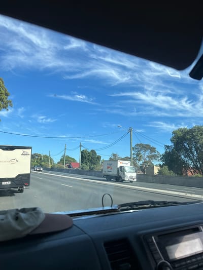 Captured from the interior of a moving vehicle, the scene presents a northbound view along a highway in Sutherland, Australia. A "Budget" rental truck is traveling in the lane to the right, and an RV is driving in the lane to the left. The sky is bright blue with thin, wispy clouds. Power lines run alongside the highway. Trees and some residential housing are visible in the background. The vehicle's dashboard and side pillar are visible in the foreground, partially obscuring the view. A white cloth or glove is resting on the dashboard. The registration plate on the RV reads "TT-36-PL".