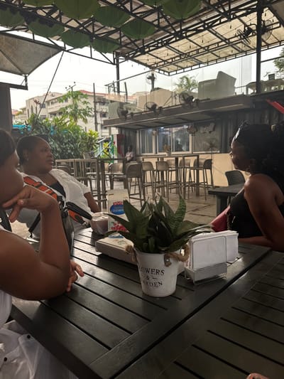 A high-angle shot shows two women at a dark wooden table, a potted snake plant in the middle, and a napkin dispenser. The women are cropped at the chest; one wears a white shirt with orange trim, the other is not visible. In the background, a restaurant interior with chairs and tables can be seen. Overhead, there's a decorative arrangement of green umbrellas. The scene is bathed in bright, natural light.