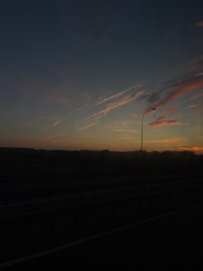 An outdoor, medium shot captures a twilight sky over a darkened landscape. The sky is a gradient of deep blue at the top to orange and yellow hues near the horizon, illuminated by wispy, pink-tinged clouds. A tall, slender street lamp with a curved arm stands silhouetted against the sky on the right side of the frame. Below the sky, a dark, silhouetted line of trees and a gentle hill forms the horizon. In the foreground, a dark, flat expanse suggests a road or highway, with faint white lines visible, indicating lanes. The overall scene is dark and atmospheric, characteristic of dusk. The location is not specified in the image.