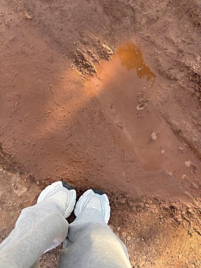 A pair of white and gray athletic shoes, worn with light gray sweatpants, are positioned at the bottom of the frame, pointing forward towards a muddy terrain. The ground is predominantly composed of reddish-brown mud, wet in places, with visible footprints and tire tracks. A shallow puddle of murky water, reflecting the sky and some indistinct foliage, is present in the upper right quadrant of the image. The lighting suggests it is either early morning or late afternoon, with warm sunlight casting long shadows across the muddy surface. The scene appears to be an outdoor, undeveloped area, possibly a trail or unpaved road.
