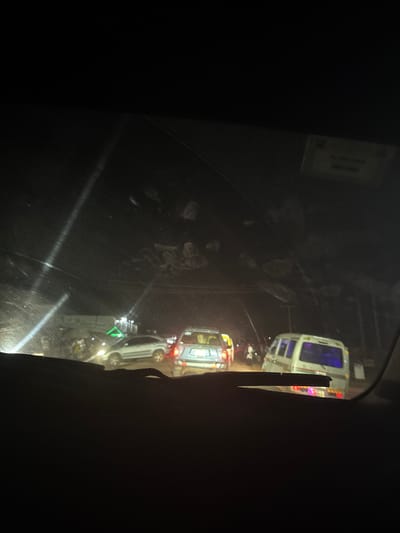 From inside a vehicle, a night scene reveals two white vans, seemingly emergency vehicles, their lights illuminated. The view is obscured by a dirty, scratched windshield. The dark surroundings suggest a nighttime setting. The vehicles are lined up, possibly at a scene requiring their attention. Streetlights or other sources of illumination create reflections on the wet or dirty surface of the windshield.