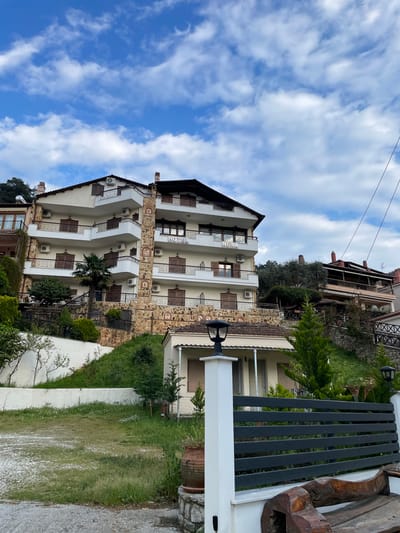 The image depicts a multi-story hotel in Skala Potamias, Greece. The hotel is white with brown window shutters and stone accents. Each level has balconies. There are air conditioning units installed on the exterior walls. The word "HOTEL" is visible on one of the upper floors. Adjacent to the hotel is a small, grassy area enclosed by a white wall. A wooden and metal fence stands near the edge of the property. In the background, other buildings and greenery are visible. The sky above is blue with scattered white clouds. Two electrical wires run diagonally across the upper part of the image.