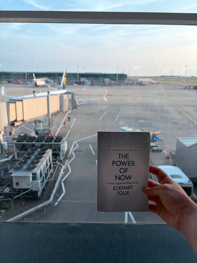 A person's hand is holding a book titled "The Power of Now" by Eckhart Tolle, subtitled "A Guide to Spiritual Enlightenment." The book cover features a stylized sunburst pattern and the text is predominantly green. The scene behind the hand and book is an airport tarmac, viewed from inside a building through a large window. Multiple airplanes, jet bridges, and ground support equipment are visible. The lighting suggests it is daytime, with the sky appearing bright and overcast. The overall impression is one of a waiting area or lounge at an airport, likely before a flight. The perspective is from above, looking down onto the tarmac.