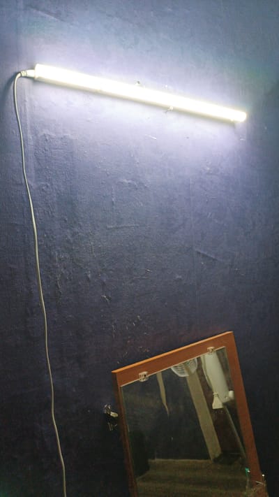 The image shows a fluorescent light fixture mounted horizontally on a dark blue, textured wall. A white power cord dangles from the left end of the light. Below and to the right of the light, a mirror with a wooden frame is propped against the wall at an angle. The mirror reflects a ceiling fan and some other indistinct objects, possibly furniture. The overall impression is of a dimly lit interior space. The location in Gryazi, Russia, is not visually indicated in the image.