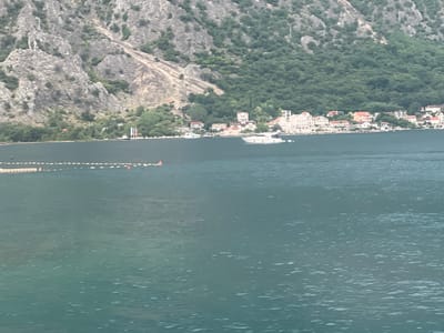 The image shows a body of water with a town and mountains in the background. The water is dark blue and appears to be calm with small ripples. In the distance, a line of small orange buoys is visible along the water's edge. Beyond the water, a cluster of buildings with red-tiled roofs and white walls forms a coastal town. Behind the town, a steep, rocky mountain rises, covered in patches of green vegetation. The sky is overcast, creating a soft, diffused light. No people are clearly visible.