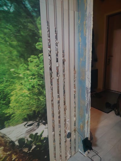 The image depicts an interior scene with a focus on a wall that is partially covered by a nature-themed mural and a vertical decorative element. The mural, on the left, displays a vibrant green forest with blurred foliage, suggesting a sense of depth. To the right of the mural is a wall composed of vertical wooden planks. Within the gaps between these planks, small, smooth, light-colored stones are visible, arranged in vertical lines. The rightmost section of the image shows a wall with a textured finish in shades of blue and gold, with visible imperfections and what appears to be unfinished plastering or paint work. Electrical wires are present, including a black power adapter plugged into a socket on the textured wall, with wires extending downwards. A sliver of a doorway and part of a room with a floor of light-colored wood-look tiles are visible in the background, suggesting a hallway or entrance area. The overall impression is of an interior space undergoing renovation or decoration, with elements of both naturalistic art and construction in progress. The location is specified as Сухарево, Russia.
