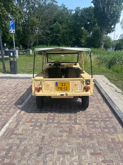 The image shows the rear view of a tan-colored, open-top utility vehicle parked on a patterned brick surface. The vehicle has a license plate that reads "'22 PB-12 NL". Behind the vehicle, there is a grassy area with tall reeds, trees, and a blue parking sign with an electric charging symbol. A dark gray charging station is visible to the left of the vehicle. The scene is outdoors on a sunny day, suggesting a location in Leiden, Netherlands, given the presence of a Dutch license plate and charging infrastructure typical of European cities.