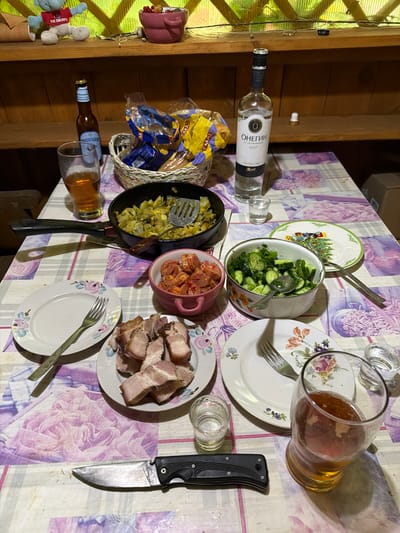 The image depicts a rustic wooden table set for a meal in what appears to be an outdoor or semi-outdoor setting, possibly a dacha in Medvedchikova, Russia. On the table, there is a bottle of vodka, a bottle of beer, a basket of snacks, a pan of cooked potatoes and vegetables, a bowl of chopped tomatoes, and a bowl of sliced cucumbers and dill salad. Two plates with slices of roasted pork are present, alongside cutlery and glasses. A knife is placed on the tablecloth. The background shows a wooden shelf with a stuffed toy and a pink bowl containing berries. The overall impression is of a casual, communal meal.