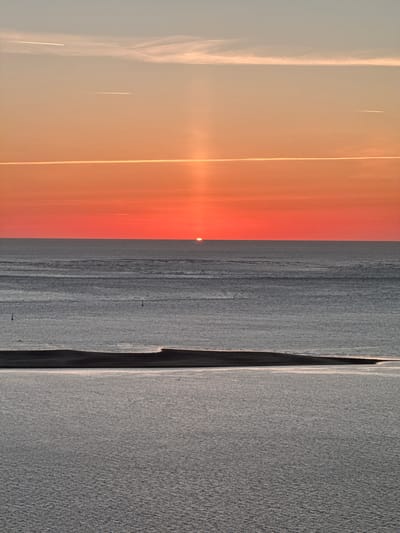 The image shows a sunset over the ocean. The sun is a small, bright disc on the horizon, casting a warm orange glow across the sky. The sky is a gradient of colors, from pale blue at the top to a deep orange-red at the horizon. There are a few contrails from airplanes visible in the sky. The ocean is calm, with gentle waves. In the foreground, there is a sandbar or coastline. The scene appears serene and peaceful. The location is likely the coast of La Teste-de-Buch, France, known for its beaches and proximity to the Atlantic Ocean.