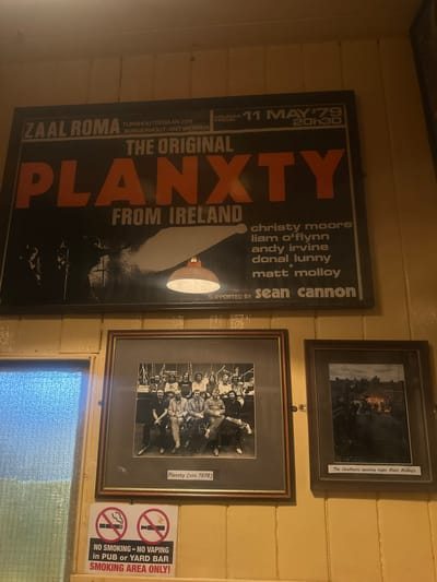 The image displays a wall decorated with framed posters and photographs, suggesting a venue related to music or performing arts. Dominating the left side is a large, dark poster for an event titled "PLANXTY - THE ORIGINAL FROM IRELAND." The poster features large, orange lettering and lists performers like Christy Moore, Liam O'Flynn, Andy Irvine, Donal Lunny, Matt Molloy, and Sean Cannon. Dates and location details are also visible: "FRIDAY 11 MAY '79 20h30" and "ZAAL ROMA TURNHOUTSEBAAN 286 BORGERHOUT-ANTWERPEN."
To the right, two framed photographs are hung. The top frame contains a black and white image with a handwritten label that reads "The Chieftains opening night Matt Molloy's." The bottom frame also holds a black and white photograph, captioned "Planxty (circ 1978)."
A "NO SMOKING - NO VAPING" sign with circular red and white prohibition symbols is prominently placed to the right of the framed photographs, indicating rules for the establishment. The overall setting appears to be indoors, with wooden paneled walls and tiled sections, possibly in a pub or bar, given the sign's text. The lighting suggests it might be evening, aligning with the concert poster's time. The atmosphere conveyed is one of historical and cultural significance, likely a place that celebrates Irish music and heritage.