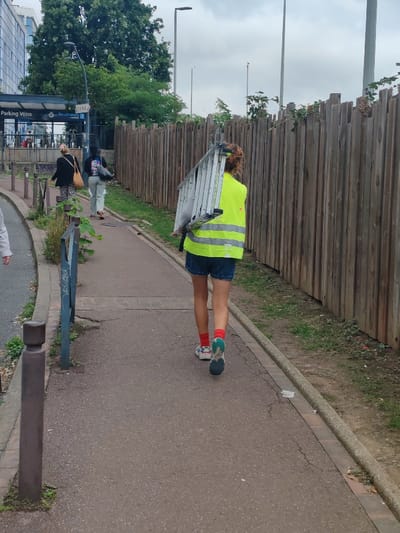 The image depicts an outdoor pedestrian pathway in Fontenay-sous-Bois, France, under an overcast sky. In the foreground, a person, viewed from behind, walks away from the camera on a reddish-brown asphalt path showing minor cracks. This individual wears a bright yellow high-visibility vest with reflective stripes, blue denim shorts, red socks, and multi-colored athletic shoes. A silver, folded two-step ladder is carried over their left shoulder.

The path is bordered on the right by a concrete curb separating it from a strip of sparse grass, which runs alongside a tall, vertical wooden slat fence extending into the background. Some small green vegetation is visible at the base of the fence. On the left side of the path, several dark cylindrical bollards and a dark metal pole are present.

Further along the pathway, two additional individuals are walking away from the camera. The person on the left wears a dark patterned top and skirt, carrying a light-colored woven bag. The person on the right wears light-colored pants and a dark top, carrying a dark shoulder bag.

In the background on the far left, a multi-story building with a glass facade is partially visible behind dense green foliage. A blue sign post reading "Parking Vélos" and displaying the "Île-de-France" logo is mounted near what appears to be a public transport entrance, indicated by visible turnstiles. Several streetlights, including a curved one over the sign and straight poles, are visible in the distance above the fence line.