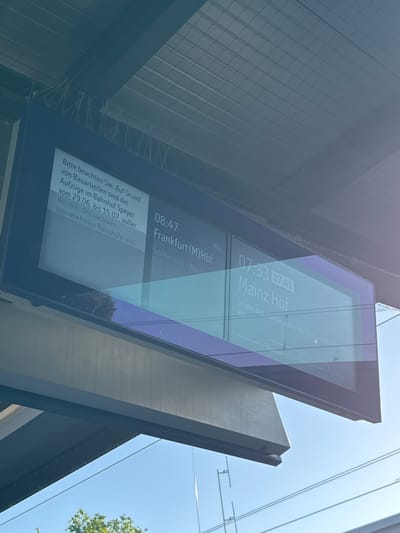 The image shows a digital train departure board at a train station, likely in Speyer, Germany. The board displays train times and destinations. One section of the board provides a message in German, stating: "Please note: Due to construction work, the elevators at Speyer train station are out of service from June 29th to July 15th. The next barrier-free train stations are...". Another section shows a departure at 08:47 for Frankfurt (M) Hbf and another departure at 07:33 for Mainz Hbf, with a listed departure time of 07:41 for the latter. The board is illuminated and reflects the surrounding environment, including the sky and overhead structures. The image is taken from a low angle, looking up towards the train station's canopy.