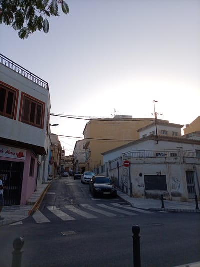 The image depicts a street scene in Olula Del Río, Spain, likely during the late afternoon or early evening due to the soft lighting and the clear sky. The perspective is from a street-level viewpoint looking uphill. The street is paved and appears to have some unevenness. A pedestrian crossing is visible in the foreground.
On the left side of the street, a building with a light-colored facade and dark wooden shutters is prominent. A pink storefront with the partial text "de Maria" is visible at the lower left. Above the storefront, a branch with green leaves hangs into the frame from the upper left corner.
Further up the street, buildings of varying heights and architectural styles line both sides. Several cars are parked or driving on the street, including a black sedan in the foreground and a white SUV further up. Power lines and streetlights are strung across the street.
On the right side of the street, a white building with a tiled roof is visible, featuring graffiti and a red circular sign with a white horizontal bar, indicating a prohibition. Another building with a yellow facade is situated behind it. A sign with the text "SE VENDE" and a phone number is attached to the white building.
Two black bollards are positioned on the sidewalk in the foreground, and another is visible on the right side of the street. The overall impression is of a residential or commercial street in a Spanish town, with signs of everyday life.