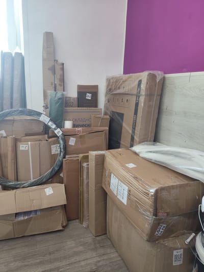 The image shows a cluttered storage area with numerous cardboard boxes stacked on a wooden floor. The boxes are of various sizes and appear to be filled with different items. Some boxes are labeled with numbers and text in Cyrillic script, such as "366," "502," and "174." One large box is wrapped in plastic and appears to contain a television, as indicated by the text "TV" visible on its surface. Another prominent object is a large, coiled black cable wrapped in plastic. In the background, a portion of a wall painted in a vibrant purple hue is visible, juxtaposed with a white wall and a window. The scene suggests a disorganized yet functional storage space, possibly a warehouse or a room used for receiving and holding inventory. The exact location within Askiz, Russia, cannot be determined from the image alone.