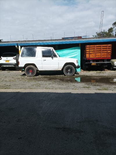 The scene depicts an outdoor parking or storage area within the city of Popayán, Colombia, under an overcast sky. In the midground, a white two-door, short-wheelbase SUV, consistent with an older model Mitsubishi Pajero, is parked facing right. This vehicle features black wheels with a red center cap on the front left wheel and a black center cap on the front right wheel. Its rear quarter windows are covered with a reflective silver film. To the left of this vehicle, a white Renault Captur SUV with a dark roof is partially visible, displaying the Colombian license plate GXP 73J on its rear hatch, which also mounts a spare tire. To the right of the white Pajero, a black truck with a wooden stake-side cargo bed is parked, displaying the Colombian license plate PEC 048. Further to the right, only the front bumper and lower fascia of a light-colored sedan or small SUV are partially visible.
All vehicles are situated on a gravel surface containing several puddles of standing water, reflecting the sky and nearby objects. In the background, a low-rise building structure with a flat, grey roof and a blue fascia runs across the frame. A light blue-green tarp or sheet is hung behind the vehicles, serving as a partition or wall. A grey corrugated downspout is visible on the left side of the building. Multiple thin, dark, pole-like structures protrude above the roofline. A more substantial metal lattice structure, possibly a small tower, is visible further in the background to the right, above the building. The foreground consists of a dark grey concrete pavement, which is partially shadowed, indicating an overhead obstruction outside the frame. No individuals are present in the scene, and no actions or interactions are depicted.