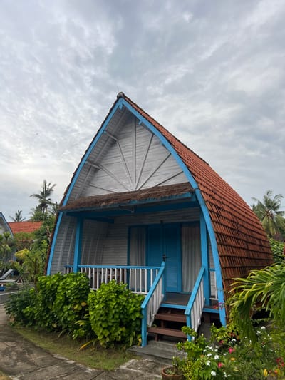 A vibrant building with a red-tiled roof and blue trim under a cloudy sky. The structure features a triangular gable end with light grey wood panelling radiating from a central point. A small porch with white railings and blue accents is present, accessed by wooden steps. Lush green vegetation surrounds the building, suggesting a tropical setting. Another building with a similar roof is visible in the lower background.