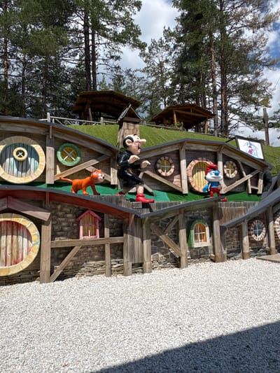 In Yurukovo, Bulgaria, a whimsical, multi-level structure resembling hobbit houses is set against a backdrop of tall pine trees and a cloudy sky. The construction features rustic wooden walls and roofs, interspersed with stone accents. Various circular windows, some with decorative crossbars, punctuate the facades.
Prominently displayed on the upper level is a large, animated figure of Gargamel, a character from "The Smurfs" cartoon, with his signature pointed nose and dark clothing. Standing next to him is an orange, cartoonish cat, also likely from "The Smurfs" series. On a lower tier, a Smurf figure, recognizable by his blue skin, white hat, and red pants, is positioned.
The surrounding ground is covered in a thick layer of white gravel. The overall scene evokes a playful, storybook atmosphere, likely part of a themed attraction or park.