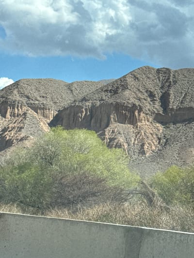 The image shows a mountain landscape under a blue sky with white clouds, near Kok Moynok Eki, Kyrgyzstan. The mountain has a rough, textured surface, and is partially covered with greenery on its right side. A gray surface, likely a road, is visible at the right edge of the frame.