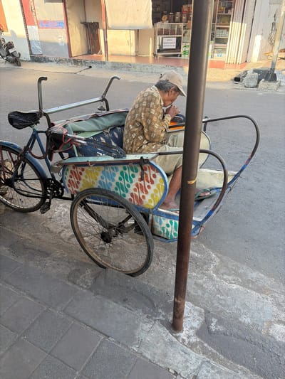This image depicts a man sitting in a pedicab on a street in Pekalongan, Indonesia. The man, wearing a cap and a patterned shirt, appears to be eating or engaged in a similar activity. The pedicab, a three-wheeled vehicle with a passenger carriage, is colorful, with a vibrant, abstract design on its side. The scene is set in an urban environment with a building in the background displaying various goods and signage, including one that reads "SEDIA BLACK GARLIC." The street surface is paved, and a motorcycle is visible parked on the left side of the frame. A metal pole stands prominently in the foreground, near the pedicab. The lighting suggests it is either early morning or late afternoon, with warm sunlight casting shadows.