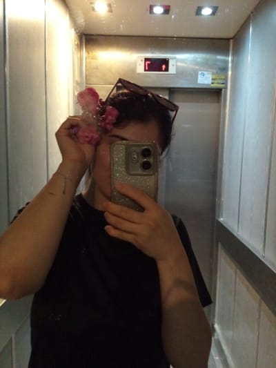 A person is taking a selfie in an elevator. They are holding a cluster of pink flowers to their ear and have sunglasses perched on their head. The elevator has brushed metal walls and a digital display showing an upward arrow and a left arrow. The lighting appears to be artificial, suggesting it is likely daytime or nighttime, and there are no windows visible to indicate weather conditions. The person is wearing a black t-shirt and a bracelet with a small charm. The scene is a typical elevator interior. No visible text on the person or the flowers. There is some text on a sticker on the elevator wall, but it is too blurry to read.