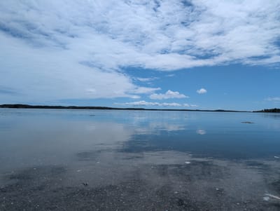This is a wide-angle, eye-level photograph of a calm body of water, likely the ocean or a large bay, with a shoreline in the foreground and a distant landmass across the water. The foreground consists of a pebbly, dark-colored surface, possibly a beach or a path near the water's edge. The water itself is clear and reflective, mirroring the blue sky and white clouds above. The sky is predominantly blue with scattered, wispy white clouds, some of which are broken and form streaks. The distant landmass appears to be covered in dark green trees and vegetation, with some hints of buildings or structures visible. The overall scene evokes a peaceful and serene natural environment. The location is specified as West Isles Parish, Canada, suggesting a coastal or island setting within this region. There are no people or animals visible in the image, and no apparent actions or interactions taking place. The objects are limited to the natural elements of water, sky, land, and pebbles.