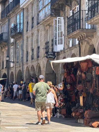 The image depicts a street scene in Santiago de Compostela, Spain, likely in the historic center near the cathedral, characterized by traditional stone buildings with arcades and balconies. In the foreground, a man wearing a green t-shirt and khaki shorts walks away from the camera, his back to the viewer. Beside him, a woman in a white tank top and white shorts walks in the same direction. Behind them, a crowd of people are seen walking along a stone-paved street, some browsing a market stall draped with numerous leather bags and accessories. The market stall is under a white awning. A sign on the stall reads "CUERO A 10€", indicating leather items priced at 10 Euros. The architecture suggests an older European city, consistent with the historic districts of Santiago de Compostela. The lighting indicates it is a sunny day.