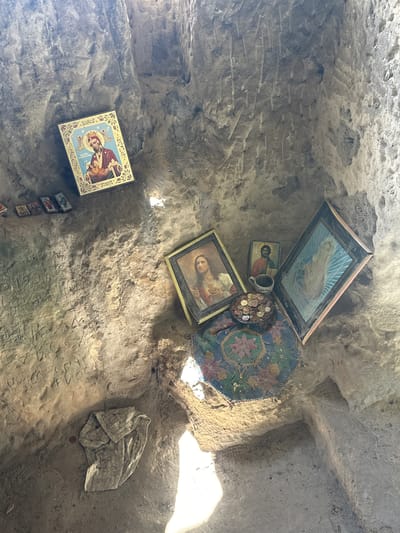 The image shows the interior of a cave or niche in Milanovo, Bulgaria. The walls are rough and appear to be stone or hardened earth. Attached to the walls are several religious icons and images in frames, of varying sizes and conditions. There is a plate or dish filled with coins placed near the icons, along with a ceramic mug. A woven rug or fabric is hung near the icons. The floor is covered with a cloth and has carved lettering or symbols on it. Sunlight enters from the right side of the frame, illuminating the interior.