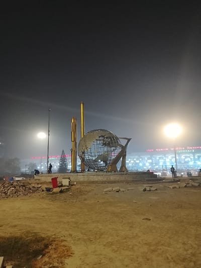 An outdoor night scene features a metallic globe sculpture surrounded by golden, rocket-shaped structures. The area appears under construction, with loose rocks and dirt covering the ground. In the background, a brightly lit building labeled "TERMINAL 2" is visible. Two individuals stand in the distance, and streetlights illuminate the scene, creating a hazy, diffused glow. The overall atmosphere suggests a place of transition or arrival.