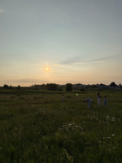 The image depicts a group of people in a grassy field at dusk. The sun is low on the horizon, casting long shadows and creating a warm, golden light across the scene. The sky is a soft blue, with wispy clouds. In the distance, there are trees and buildings, suggesting a rural or suburban setting.

Several individuals are scattered throughout the field, engaged in what appears to be some form of movement or dance. One person is bent over, as if stretching or reaching for the ground. Another is in mid-air, possibly jumping or flipping. Their clothing is casual, suggesting an outdoor recreational activity. A small, round, green object, possibly a ball, is visible in the grass between two of the individuals. The overall mood is serene and peaceful, enhanced by the natural beauty of the landscape and the soft lighting.

The location context is Kutlu-Bukash, Russia. The time of day is clearly evening, given the position of the sun. The weather appears to be clear and calm. There is no visible text in the image.