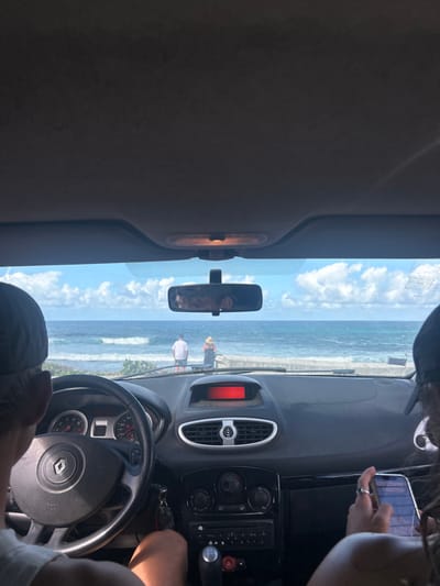 The image provides a forensic view from inside a Renault vehicle, looking out towards a coastal scene in Le Moule, France.
In the foreground, the car's interior is visible. On the left, the driver, wearing a dark cap, has their left hand on the black steering wheel, which bears a silver Renault logo. The dashboard features a red digital display, air vents, and climate controls. A manual gear stick is visible, with keys hanging from the ignition. On the right, a passenger, also wearing a dark cap, holds an illuminated smartphone in their right hand. The rearview mirror reflects two indistinct figures, likely the occupants. An overhead interior light is visible on the ceiling.
Beyond the windshield, the Atlantic Ocean with moderate waves extends under a blue sky dotted with scattered white clouds. A red buoy and a white buoy are visible in the water. On the immediate shoreline, behind a low barrier, two individuals are standing and observing the sea. The person on the left is a male, seen from behind, dressed in a white t-shirt and white shorts. The person on the right is a female, also viewed from behind, wearing a dark patterned dress and a wide-brimmed straw hat.