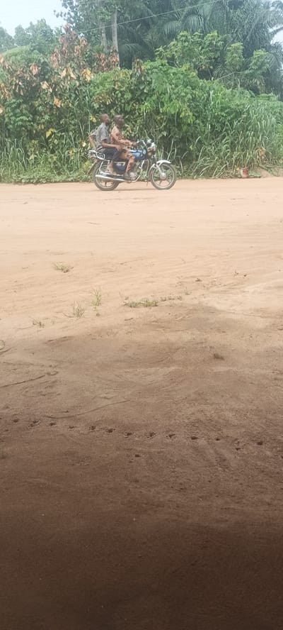 Two men are riding a blue motorcycle on a dirt road in Amauzu, Nigeria. The road is lined with dense vegetation. The foreground is dusty and partially shadowed. There are animal tracks in the foreground.