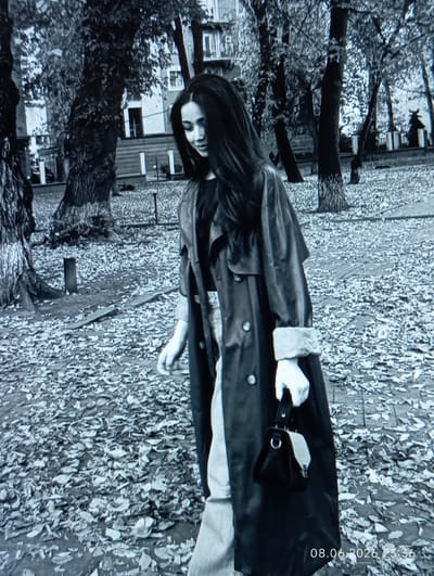 A young woman with long dark hair, wearing a trench coat and light-colored pants, walks through a park covered in fallen leaves. She is carrying a dark handbag. The background shows trees and buildings. The image has a timestamp in the lower right corner indicating June 8, 2026, at 23:36.