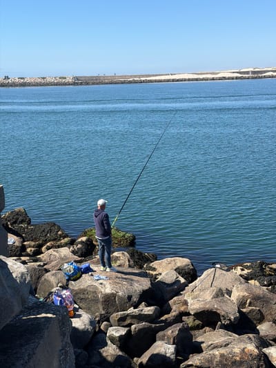 The image depicts a bright, sunny day at the Gafanha da Nazaré marina in Portugal, specifically overlooking the mouth of the Aveiro Lagoon. In the foreground, a man wearing a blue hooded sweatshirt, jeans, and a white baseball cap stands on a rocky shore, fishing with a long rod. He is facing away from the camera, towards the calm, blue water.
Scattered among the rocks are several bags, including a blue shopping bag and a bag with a pink and white pattern. A white cloth is also visible on one of the rocks. Further out in the water, a long, concrete breakwater extends into the sea, with sand dunes visible beyond it. A few people can be seen walking along the breakwater in the distance. The overall scene is one of peaceful, outdoor recreation by the water.