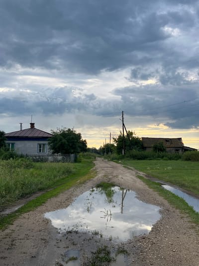 The image depicts a rural landscape in Pivkino, Russia, likely during the late afternoon or early evening, given the warm, soft light filtering through overcast skies. The scene is dominated by a dirt road that curves into the distance, flanked by overgrown grass and vegetation. A large puddle reflects the dramatic, cloudy sky and the surrounding environment, adding a sense of depth and mirroring.
On the left side of the road, a modest house with a dark-colored roof and light-colored siding is visible, bordered by a simple fence and dense greenery. Further in the distance, the silhouette of a partially ruined building, possibly an abandoned structure or barn, stands against the sky, hinting at the passage of time and decay. Utility poles with power lines traverse the sky, indicating the presence of infrastructure even in this seemingly remote setting.
The weather appears to be partly cloudy, with heavy, dark clouds in the upper left portion of the frame gradually giving way to brighter, more illuminated patches of sky near the horizon. This contrast suggests a dynamic weather system, perhaps a storm passing or approaching, which contributes to the somber yet beautiful mood of the photograph. The overall impression is one of quiet rural life, touched by elements of nature's power and the signs of human habitation, both present and past. There is no visible text in the image.