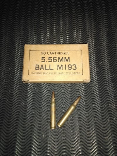 This image, located in the city of Tas-Kop', Kazakhstan, depicts a forensic scene featuring ammunition components. Dominating the upper center is an open, light-brown cardboard box labeled "20 CARTRIDGES", "5.56MM", "BALL M193", and with a "WARNING: KEEP OUT OF REACH OF CHILDREN". Positioned directly below the box are two individual 5.56mm M193 cartridges. Both cartridges are metallic, exhibiting brass casings and copper-colored projectile tips. One cartridge is oriented vertically, while the other lies diagonally to its right. All items rest on a dark, textured surface characterized by a repetitive wavy or ridged pattern. The lighting is low, accentuating the details of the objects against the background. No people are present.