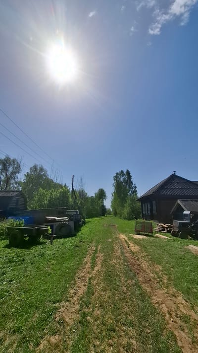 A daytime landscape photograph taken in Sheshurga, Russia, shows a rural scene. The sun is bright in the upper left, shining across a blue sky with some clouds. To the right, a dirt path with tire tracks runs from foreground to background, bordered by green grass. An old wooden building is visible at the top right. A tractor is parked near the center-right of the image. Trees are visible in the mid-ground. Power lines run across the lower left of the image.