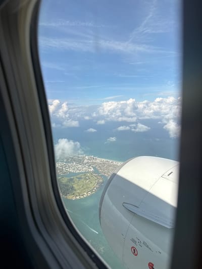 View from an airplane window over North Bay Village, Florida, USA. The airplane's engine nacelle is visible in the upper right quadrant. Below the engine, an island with a golf course and surrounding residential buildings is seen. The Atlantic Ocean extends beyond the island, with scattered clouds visible in the sky. The sky transitions from clear blue to hazy near the horizon. A small boat is visible in the water near the island. The window frame is visible in the foreground.