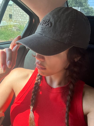 A woman wearing a red sleeveless top and a dark baseball cap with a Fila logo is shown from the chest up, sitting in a car. Her hand, with long, light-colored fingernails, is raised to adjust the brim of the cap, obscuring her eyes. Her lips, with a glossy finish, are slightly parted. Two braided brown pigtails rest on her shoulders. The background visible through the car window suggests a rural or suburban setting with a stone building and green foliage, potentially in Byurakan, Armenia. The sunlight casts strong shadows and highlights on the woman's skin and clothing.