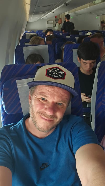The image shows a man taking a selfie inside an airplane cabin. He is wearing a blue t-shirt and a baseball cap with a "DARK SEAS" logo. His face is partially visible, and he is smiling. In the background, other passengers are seated in blue airplane seats. Some passengers are looking at their phones or resting. The airplane appears to be on the ground or in the air, and the lighting suggests daytime. The location is identified as Bao'an District, China.