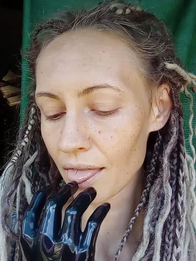 A close-up portrait depicts a woman with her eyes closed or cast downwards. Her facial skin displays a natural complexion with scattered freckles. Her hair is composed of numerous dreadlocks, presenting a blend of dark brown and lighter grey/blonde strands. Her lips are light pink and slightly parted, with her tongue gently touching the glossy black fingers of an anatomically shaped prosthetic hand. The hand is highly reflective, indicative of a hard, polished material. The background is predominantly a soft, out-of-focus deep green, likely fabric or vegetation, with a darker, indistinct area featuring horizontal elements visible on the left. The scene is illuminated by natural light and is located in Ko Siboya, Thailand.
