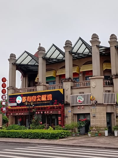 The image captures a multi-story building in Shuangfu, China, under an overcast sky. The building features an elaborate architectural style with prominent beige columns, each topped with a decorative finial or dome. Between the columns are glass-paneled awnings or skylights, and below them, yellow and orange fabric awnings extend over storefronts on the ground floor.

The ground floor of the building houses what appears to be a restaurant or commercial establishment. A large, illuminated black sign, trimmed with yellow, prominently displays Chinese characters 梁家饺子 (Liángjiā Jiǎozi), which translates to Liangjia Dumplings, along with its Pinyin transliteration "LIANGJIAJIAOZI." The sign also features a circular logo depicting a red and green stylized bird or phoenix. Below this main sign, several smaller red circular signs with white characters are visible; one clearly reads 招牌 (Zhāopái), indicating a signature dish or brand. Further to the right, a rectangular sign with black and white horizontal stripes also carries the same red and green logo.

The building's lower section is adorned with additional decorative elements, including red railings, possibly representing fences or lanterns, and lush green hedges and potted plants lining the sidewalk. The sidewalk itself is a paved area with distinctive white zebra crossing stripes on the far right, indicating a street crossing. There are no people visible, suggesting a quiet moment. The overall impression is that of a bustling commercial area, temporarily devoid of activity, possibly during a cloudy day.