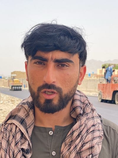 A man stands outdoors in what appears to be قندهار, Afghanistan, possibly during road construction or repair. He has dark hair and a beard, is wearing a brown and white patterned scarf and a dark green button-down shirt. Behind him are pieces of road construction machinery. A steam roller is visible in the distance, and closer, a flatbed motorized vehicle holds a worker wearing a blue shirt who is standing on what appears to be green shrubbery. The sky is overcast, and there are low-lying buildings and mountains visible in the background.