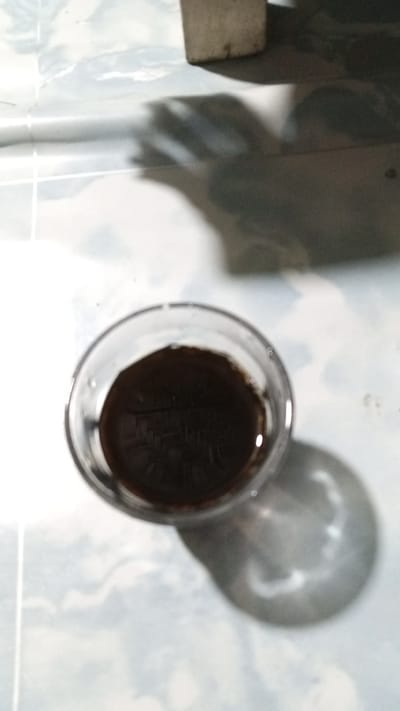 A clear glass contains a dark liquid, possibly coffee, and sits on a patterned floor. The glass casts a shadow on the floor. In the background, a light-colored object casts a larger shadow. The scene is likely indoors, with tiled flooring. The photograph was taken in Uteunreutoh, Indonesia.