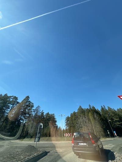 The image depicts a road in Plieņciems, Latvia on a clear day. A black Volvo station wagon is partially visible on the right side of the road with license plate NK-3649. Road signs and trees line the right side of the road. The sky is blue with a contrail from an airplane visible on the left side. The perspective is from inside a vehicle.